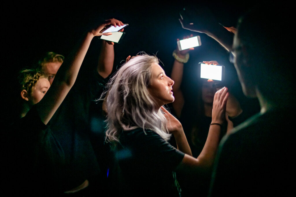 A girl wearing black is lit by the light of mobile phone screens being held up around her.