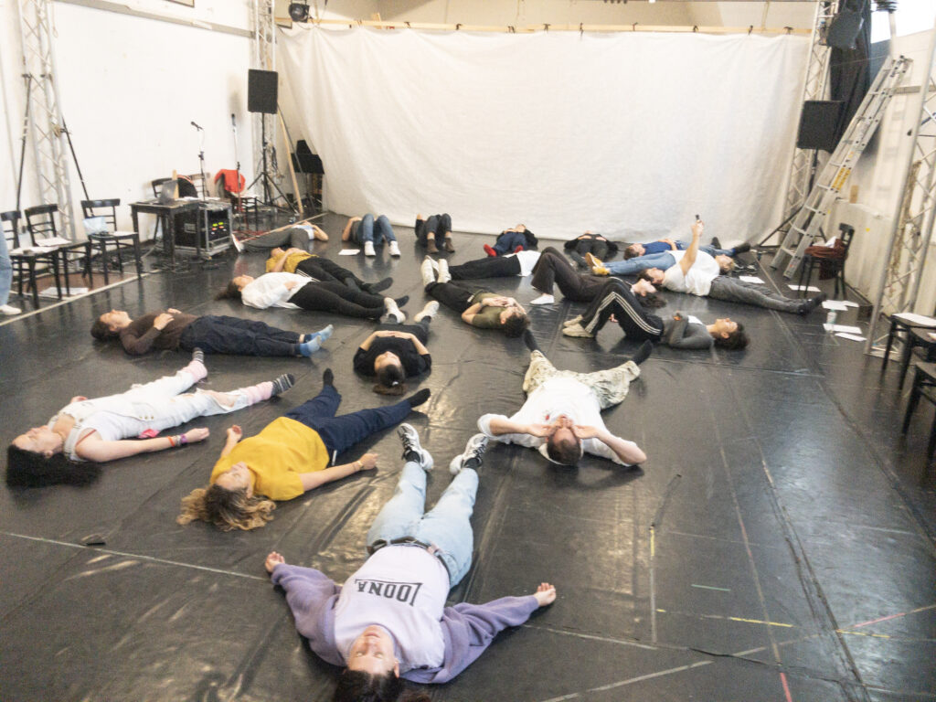 The lab participants laying on the floor during a workshop. They are in a white rehearsal studio with a black dancefloor.