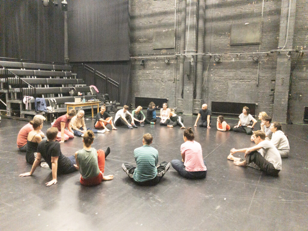 The group sitting in a circle in a black box theatre space with brick wall and seating bank behind them.