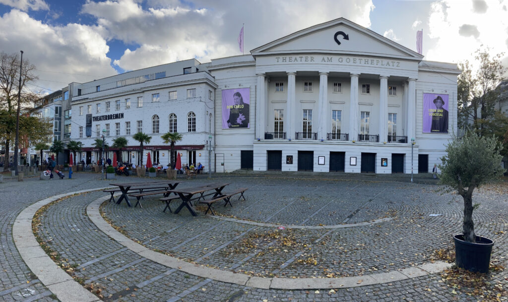 The front of the Theater Bremen main stage, a white building with columns at the front and the Theater Bremen Logo of an arrow.
