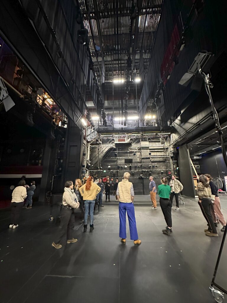A group of people on a tour of the theatre. they are on the main stage and you can see the height of the fly tower above them