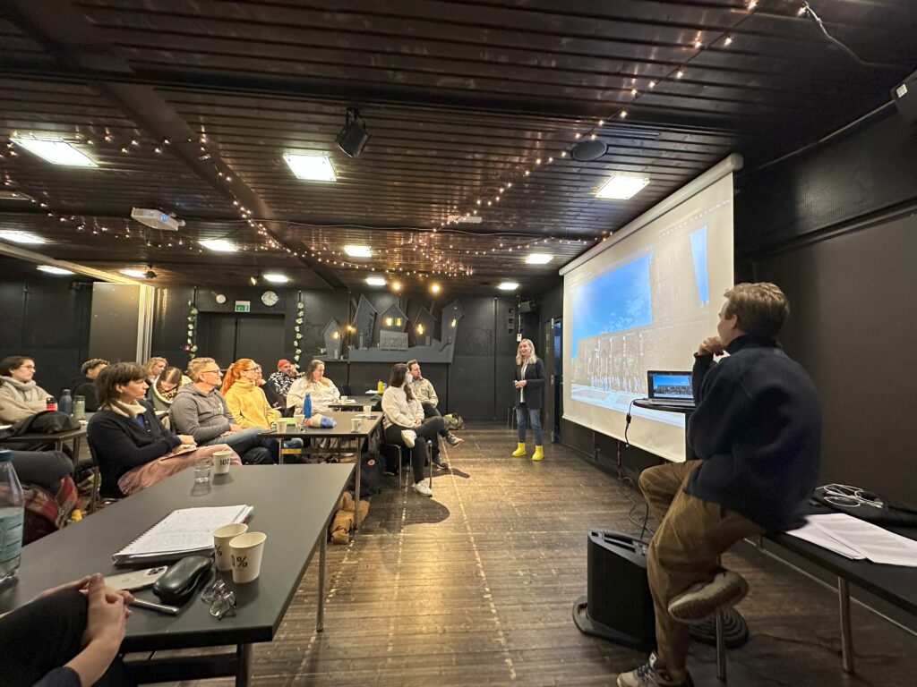 A group of people sit at tables looking towards a person making a speech in front of a screen at the front of the room.