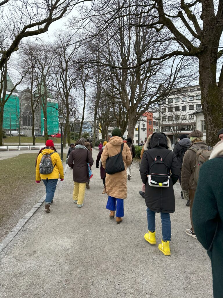 A group walk through a park in the centre of Stavanger on a winter day. They all have hats or hoods and winter books and the ground has frost on it.